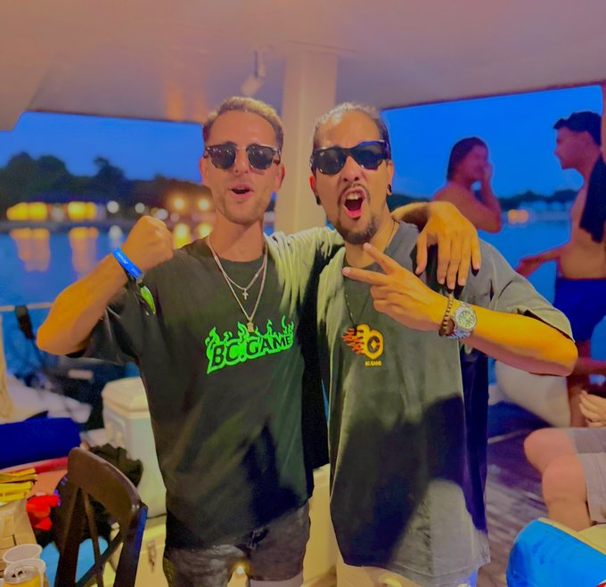 Two guys in sunglasses pose excitedly at what appears to be a beachside or poolside venue, with one wearing a shirt reading "BC.GAME" and the other wearing an "FC BARCELONA" logo shirt, while other patrons and twinkling waterfront lights fill the background — fitting the casual, celebratory "Gn Fam" (goodnight family) vibe of the post.