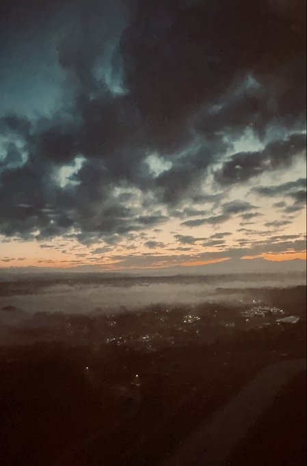 A scenic photograph of a sunset over a misty valley with a town visible below, featuring dramatic dark clouds and an orange horizon.