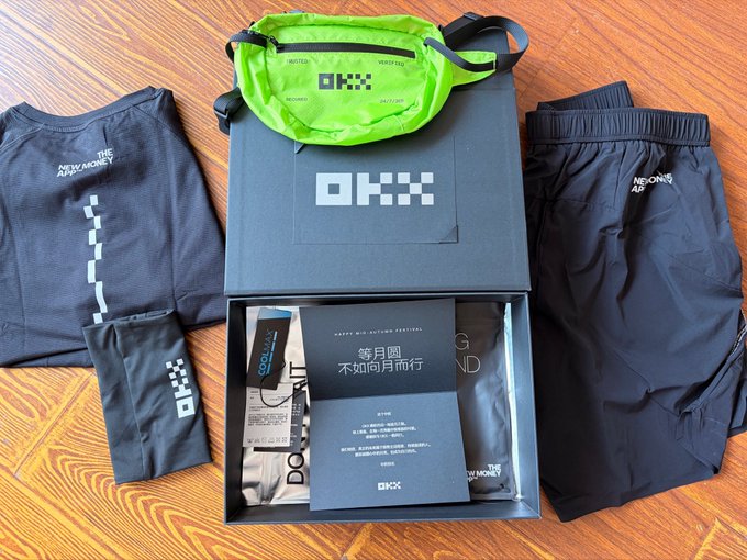 A flat lay showcasing an OKX (OKXX) Mid-Autumn Festival gift box containing a neon green crossbody bag, a black athletic shirt, black shorts, and a wrist wallet, alongside Chinese text reading "等月圆 不如向月而行" (a play on words meaning "Wait for the full moon, or walk toward the moon"), celebrating the holiday with branded apparel and accessories.