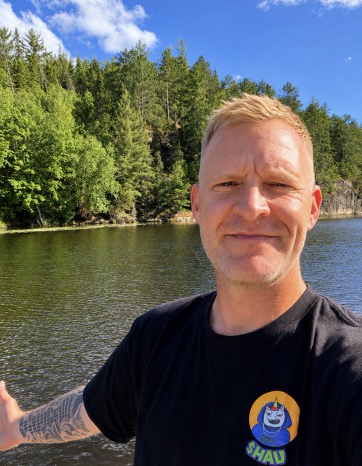 The image features a man wearing a black t-shirt with a cartoon character on it, standing by a body of water near some trees. The man has blonde hair and appears to be smiling for the camera. There are also tattoos visible on his arm. In the background, there is a post that reads "Touching some grass this week. Keep supporting each other." This message seems to be related to the image or the context surrounding it.