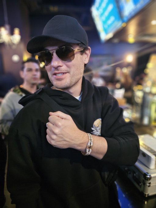 A man wearing a black cap, sunglasses, and black hoodie poses confidently in a lounge environment, gripping his shirt fabric.