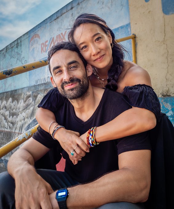 A smiling couple sits closely together on stairs, with the woman embracing the man from behind, set against a wall featuring a partially visible "RE/MAX" logo. The image appears to accompany the post's personal announcement, possibly depicting the author with his partner as he shares the difficult news about canceling his Kickstarter project.