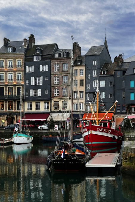 A picturesque harbor scene features colorful European-style buildings in the background with sailboats and a red fishing boat docked in the calm water.