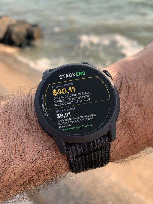 A close-up of a Coros smartwatch on a wrist at the beach, displaying a 'STACKERS' app screen with 'TOTAL EARNED $40.11' and 'IN THE VAULT $6.91' alongside various crypto tickers.
