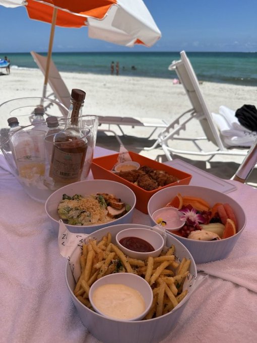 A beachside meal spread features crispy fries with dipping sauces, a colorful fruit bowl with an orchid garnish, a salad with greens and tomatoes, and fried chicken in a red container, paired with a bottle of rum and drinks on an ice-filled bucket under an orange and white umbrella. The "GM" (Good Morning) post perfectly complements this idyllic seaside brunch scene.