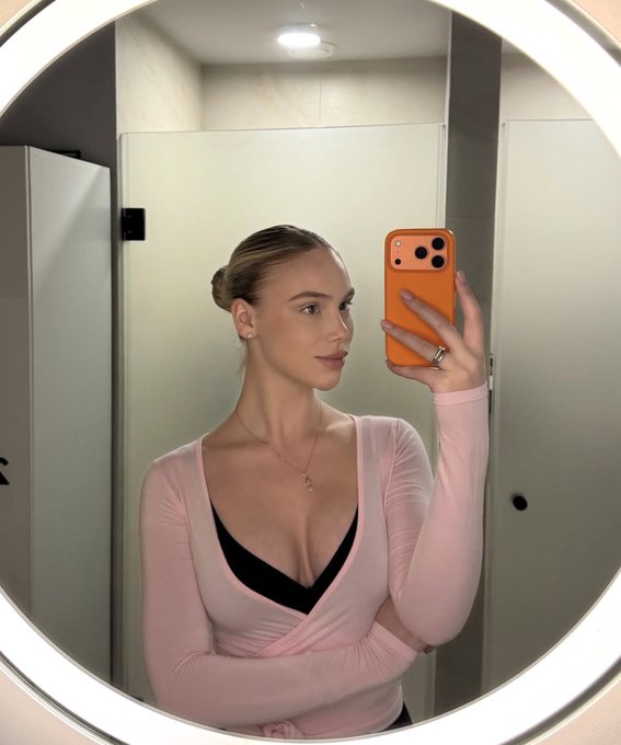 A woman with blonde hair in a bun takes a mirror selfie wearing a pink wrap top and black bra, holding an orange smartphone in a bathroom.
