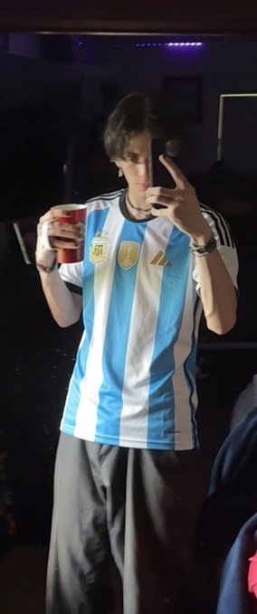 A young man takes a mirror selfie wearing an Argentina national team jersey with three stars, holding a red can.