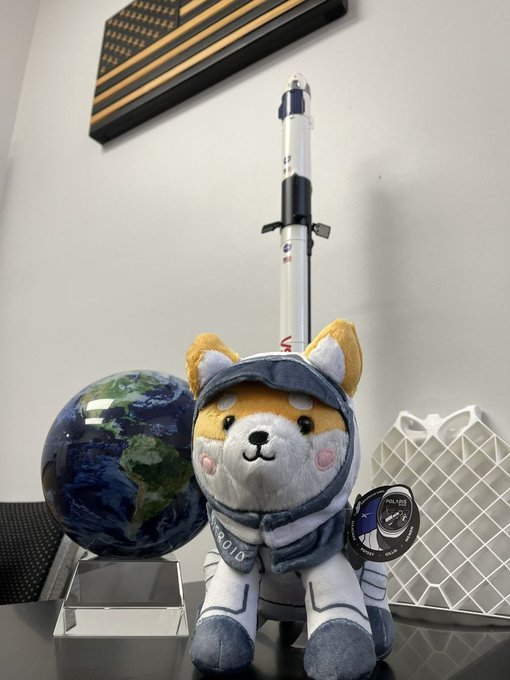 A plush toy of an orange and white fox wearing a gray astronaut suit, sitting next to a model rocket, a decorative globe, and an American flag in a display case.