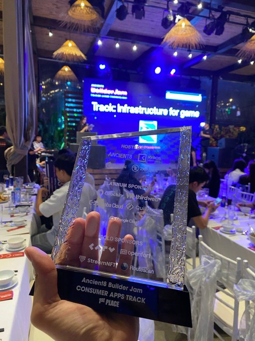 The image shows a person holding up a trophy with a plaque that reads "Ancient builders jam". The trophy appears to be made of glass, and there are several people sitting at tables around them. The background features a stage or performance area with lights on, suggesting that this event might be related to a competition or performance.
