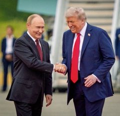 Two men in suits—one in a dark suit with red tie, the other in a blue suit with red tie—shake hands and smile at each other outdoors.