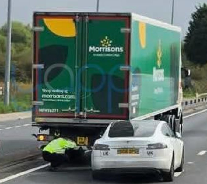 A white sports car sits wedged underneath the back of a green Morrisons delivery truck on a highway, with an emergency responder in a high-visibility jacket attending to the scene.