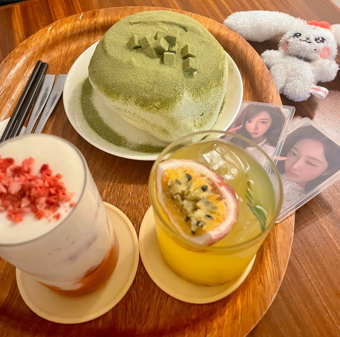 A wooden tray holds a slice of green matcha mille crepe cake, a white plush bunny, two photocards of a person, and two beverages including a layered drink topped with freeze-dried strawberries and a passion fruit drink.