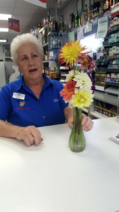 An older woman with white hair wearing a blue uniform shirt standing behind a white counter in a liquor store, next to a vase of colorful flowers.