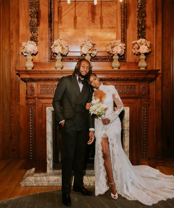 A couple poses in front of an ornate wooden fireplace mantel decorated with flowers; the man wears a black tuxedo, and the woman wears a long-sleeved white lace wedding dress with a thigh-high slit.