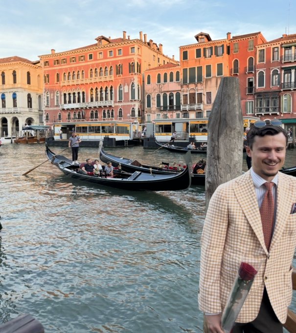 A man in a checkered blazer and tie stands in Venice, Italy, holding a red rose, with gondolas and colorful historic buildings visible on the canal behind him.