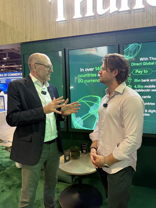 Two men in business casual attire converse at a NuPay booth, with one gesturing while speaking. The display behind them highlights features like 'Pay to' in over 140 countries and 90 currencies.