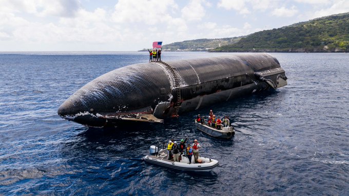 A capsized submarine floats partially submerged in deep blue ocean water near a mountainous coastline, with an American flag displayed and small rescue boats with personnel gathered nearby.
