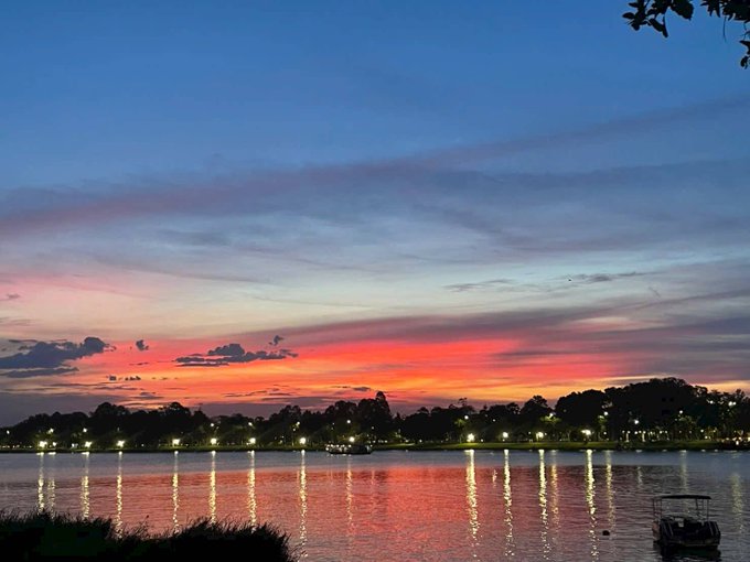 A vibrant sunset paints the sky in fiery hues of pink and orange over a tranquil lake, where lights from the distant shore reflect across the calm water like glowing promises. The serene beauty of this scene embodies the quote's spirit—just as the sky freely displays its colors at day's end, finding joy in this peaceful moment brings a deep, unhurried happiness.