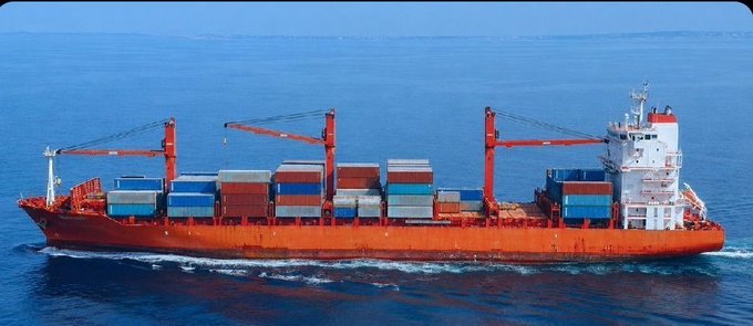 A large red cargo ship loaded with shipping containers sails across blue ocean waters, featuring tall cranes and a white bridge structure.