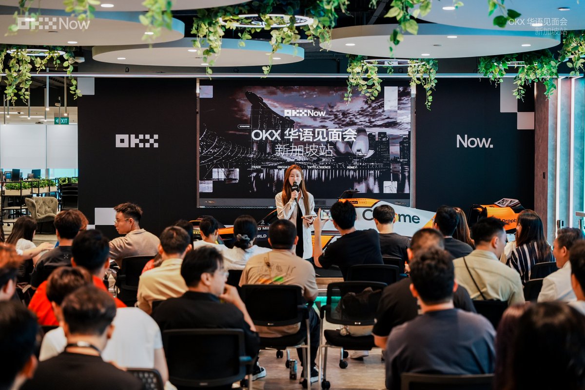 A female speaker presents at the "OKX 华语见面会 新加坡站" (OKX Chinese Community Meetup - Singapore) event, addressing a packed audience in OKX's Singapore headquarters, where an F1 race car is parked to the side as mentioned in the post.