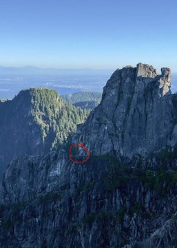 A scenic mountain landscape showing a prominent rocky peak, a forested ridge, and a distant sea or lake under a clear blue sky, with a small area circled in red on a steep rocky face.