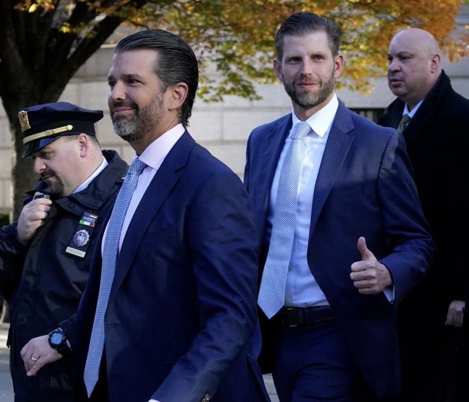 Two well-dressed men in dark suits walk alongside uniformed security personnel and another suited individual, with one man giving a thumbs-up gesture as they pass by autumn foliage and a stone building.

The image appears to show Donald Trump Jr. and Eric Trump, whose involvement with World Liberty Financial ($WLFI) — the Trump family crypto venture referenced in the post — is relevant given the company's pursuit of a federal banking charter and its intersection with the Clarity Act ethics debate.