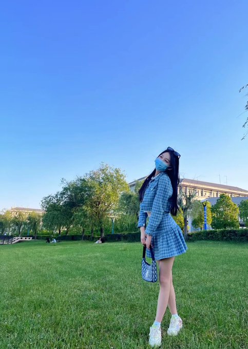 A young woman in a blue checkered outfit and mask poses on a sunny grassy field, looking up at the clear sky—reflecting the post's contrast between her past academic life and her current free-spirited crypto career, celebrating the World Cup with Bitget.
