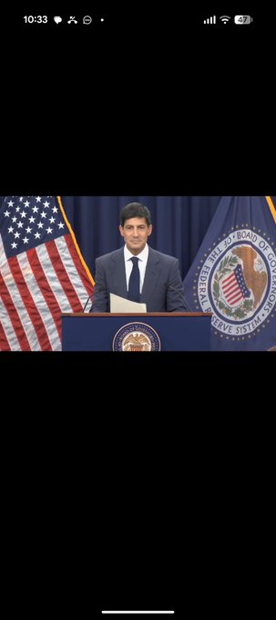 A screenshot of a phone showing a man in a dark suit speaking at a podium flanked by American flags and the seal of the President's Intelligence Advisory Board and Science and Technology, with the phone's status bar showing 10:33.
