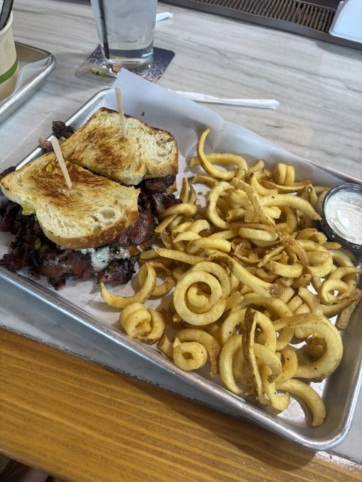 A metal tray holding a large, toasted sandwich secured with toothpicks, served alongside a generous portion of curly fries and a small cup of dipping sauce on a light wooden table.