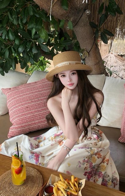 A woman wearing a 'Christian Dior' straw hat and a floral strapless dress poses at an outdoor table with a tropical drink and fries.