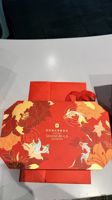 An ornate red gift box with gold fish and floral designs from Hotel Shangri-La Shenzhen, accompanied by a matching red gift bag and ribbon on a white table.