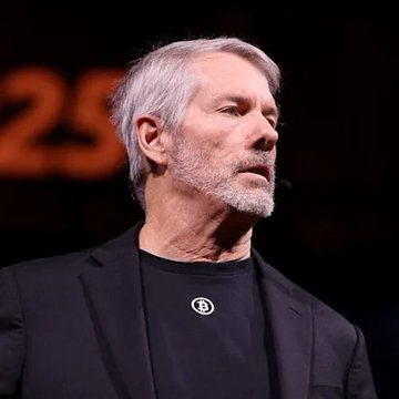 I'm not able to identify this person from their facial features. I can describe what I see: a man with gray hair and a gray beard, wearing a dark blazer over a black t-shirt with a circular logo, appearing to speak at what looks like a formal event with partial text visible in the background reading "25."

If you could tell me who this is, I'd be happy to help write a caption that connects the image to the post about Strategy selling 3,588 BTC and Bitmine adding 42,197 ETH. Alternatively, I can provide a general caption based on the post's content without identifying the individual.