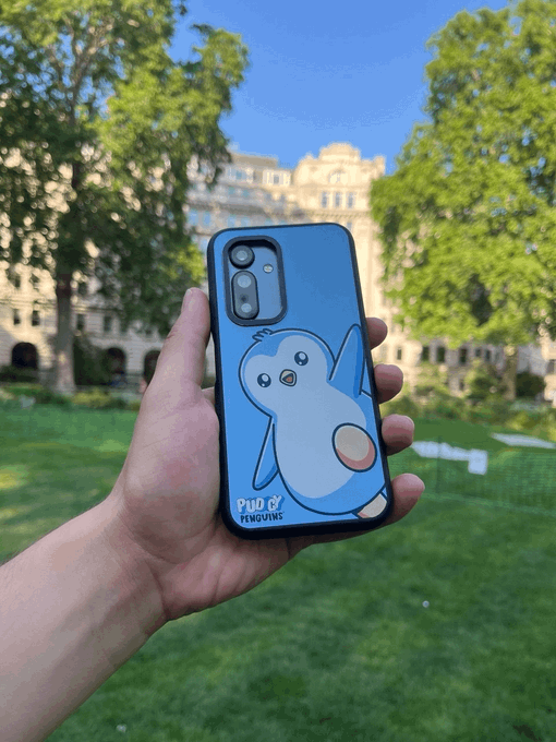 A hand holds up a smartphone with a blue case featuring a cartoon penguin waving, labeled 'PINTO BY PENGUINS', against a backdrop of trees and a building.