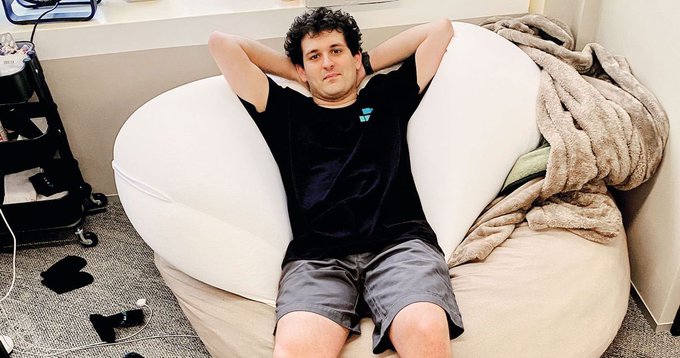 A young man with curly hair relaxes on a large white bean bag chair with his hands behind his head, wearing a black t-shirt and gray shorts.