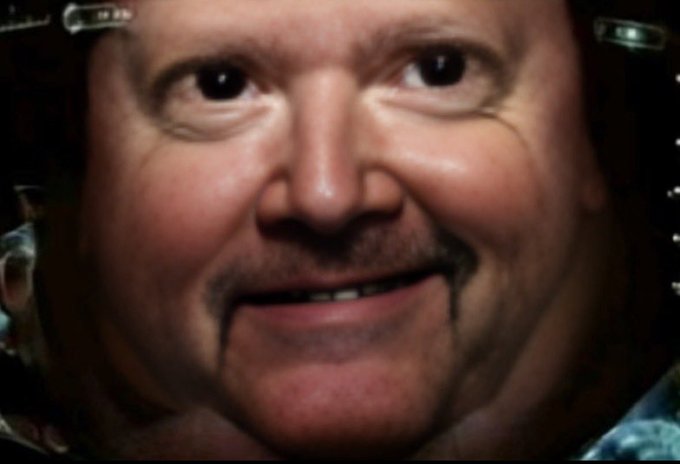 A close-up of a man's face with digitally altered features, showing an unnaturally wide smile and intense stare, resembling the character Matrick.