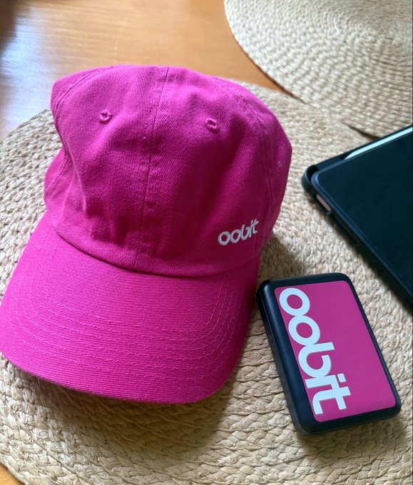 A bright pink baseball cap with the "ooht" logo embroidered on the front sits on a woven straw placemat next to a black case featuring the same "ooht" branding in matching pink, with a smartphone visible in the corner.