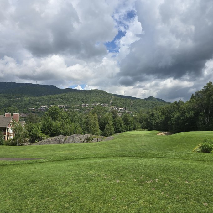 A scenic photograph of a lush green golf course fairway with rolling hills, mountains, and a cloudy blue sky in the background.