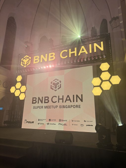 A grand stage setup at the BNB Chain Super Meetup Singapore features a large LED screen displaying the event's logo alongside sponsor names like FDUR, Aster, Brevis, and ListaDAO, with the iconic yellow hexagon branding prominently illuminated. The atmospheric venue with dramatic lighting captures the scale of the crypto event described in the post, where crowds gathered for the highly anticipated meetup.