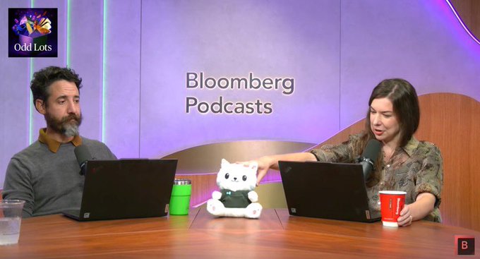 Two podcast hosts at a Bloomberg Podcasts desk with a small white cat plushie between them, with an 'Odd Lots' logo in the corner.
