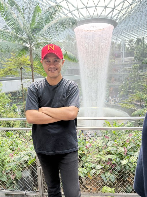 A smiling man wearing a gray t-shirt and a red cap with a yellow 'M' logo stands with crossed arms in front of a large indoor waterfall surrounded by tropical plants, resembling the Jewel Changi Airport.