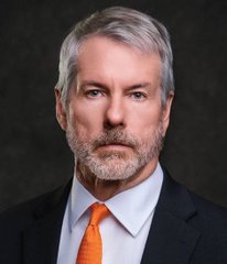 A professional headshot of a middle-aged man with gray hair and a beard, wearing a dark suit, white shirt, and orange tie, against a dark background. The image appears to be unrelated to the post's content about $LUNC cryptocurrency heading toward $0.1 and Bitcoin.