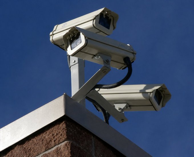 Two beige surveillance cameras mounted on a metal bracket atop a brick pillar, angled outward against a clear blue sky.