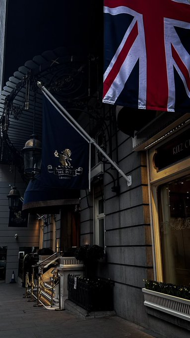 A dark nighttime photo of The Ritz London hotel entrance, featuring its iconic Union Jack flag and a navy blue flag with the hotel's crest and lion emblem, alongside gold-trimmed doors and an ornate awning.