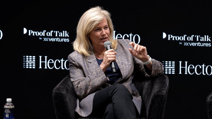 The image features a woman sitting in a chair with her hands raised while holding a microphone. She appears to be speaking into it or gesturing towards something off-camera. The background of the image includes text that reads "ProfitOfTalk", "Hecto", and "Your funds, your freedom." This suggests that she is addressing an audience at a conference or event related to financial matters or economic policies.