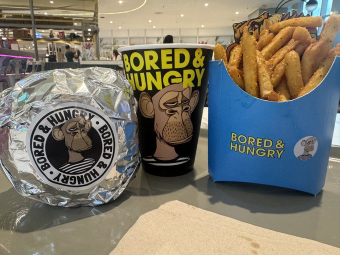 A meal from 'Bored & Hungry' featuring a foil-wrapped burger, a branded cup, and a blue carton of seasoned fries.