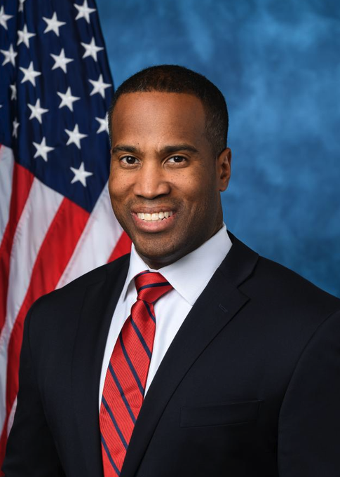 A smiling man in a dark suit with a red striped tie stands in front of an American flag. The image appears to be an official portrait of Rep. John James, who Polymarket projects has secured the Republican nomination for Michigan governor with greater than 99% probability.