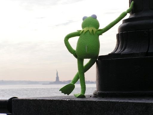 A green Kermit the Frog figurine leans against a dark lamppost on a waterfront ledge, with the Statue of Liberty visible across the water under an overcast sky.