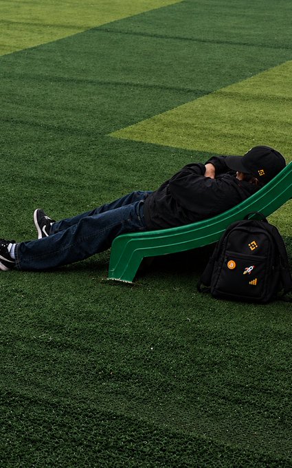 A man in a black cap and jacket lies on a green curved bench beside a black backpack on a striped artificial-turf field. The cap and backpack display orange/yellow logos or symbols, while the post’s “Strategic pause. Position held.” suggests he is deliberately resting or waiting in position.