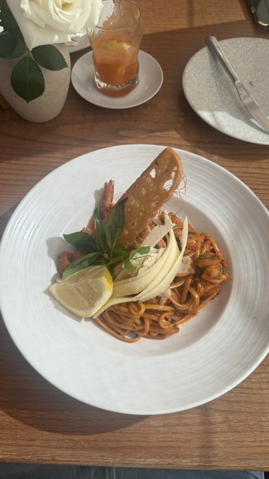 A plated dish of spaghetti with red sauce, garnished with lemon slice, fennel, basil, and a tuile crisp, served at a restaurant table with white roses and a drink in the background.