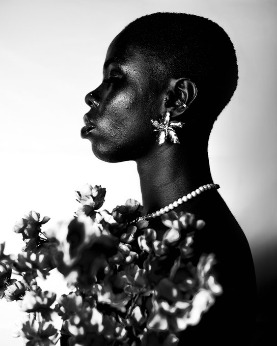 A striking black-and-white profile portrait of a Black woman with a short haircut, wearing large flower-shaped earrings and a pearl necklace, with white flowers in the foreground.